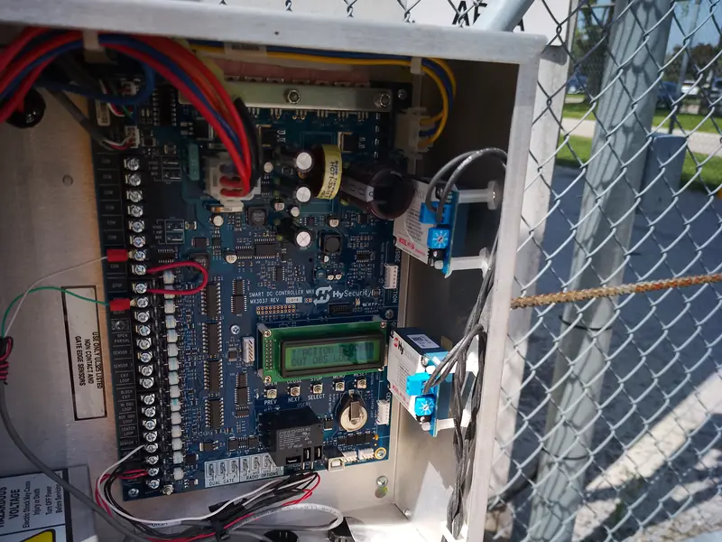 Open control box of a gate operator beside a chain-link gate.