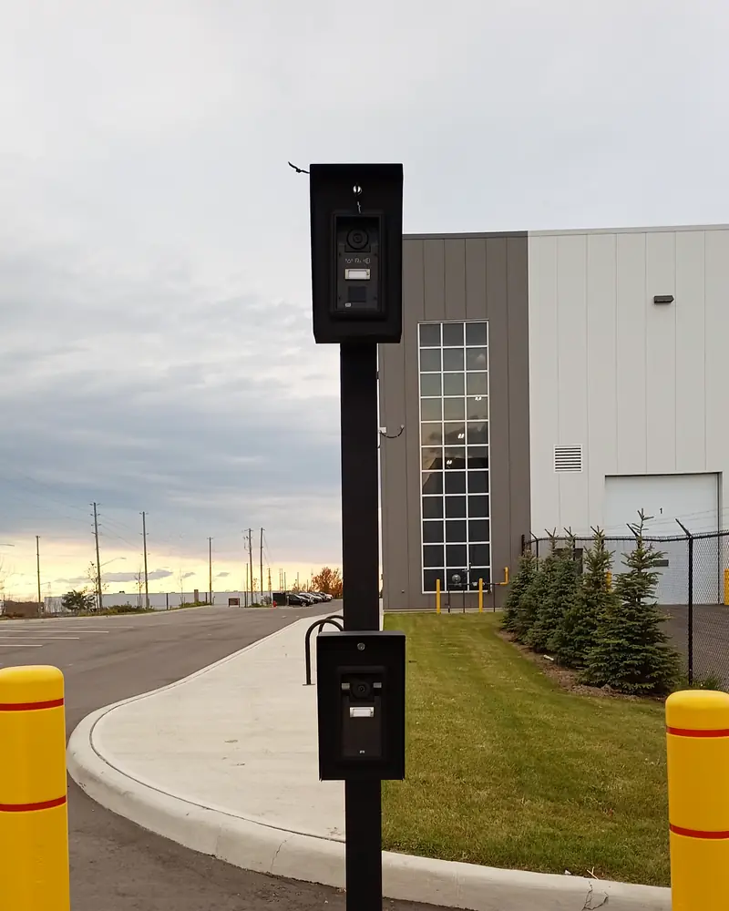 Pedestal at a vehicle entrance with intercom stations at car and truck height, between two bollards.