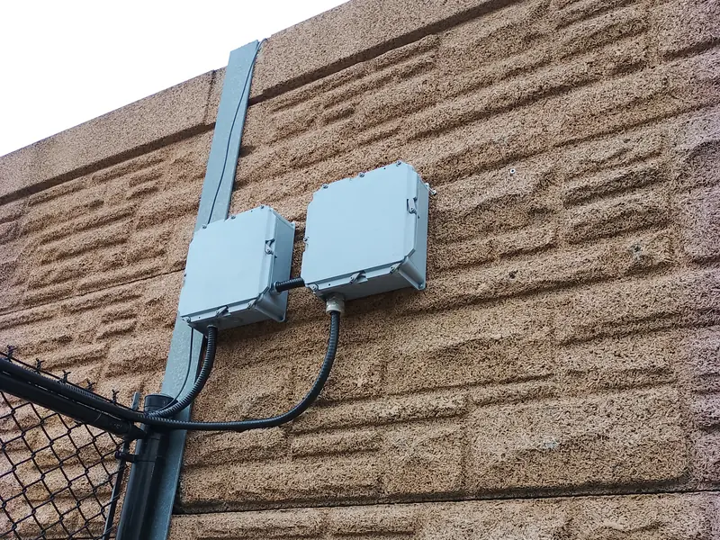 Two weatherproof junction boxes on a stone wall with flexible conduit to a fence post.