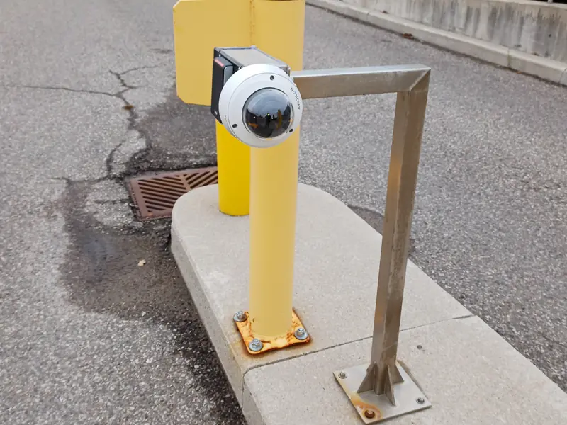 Dome camera on a steel arm bolted to a yellow bollard at a vehicle lane.